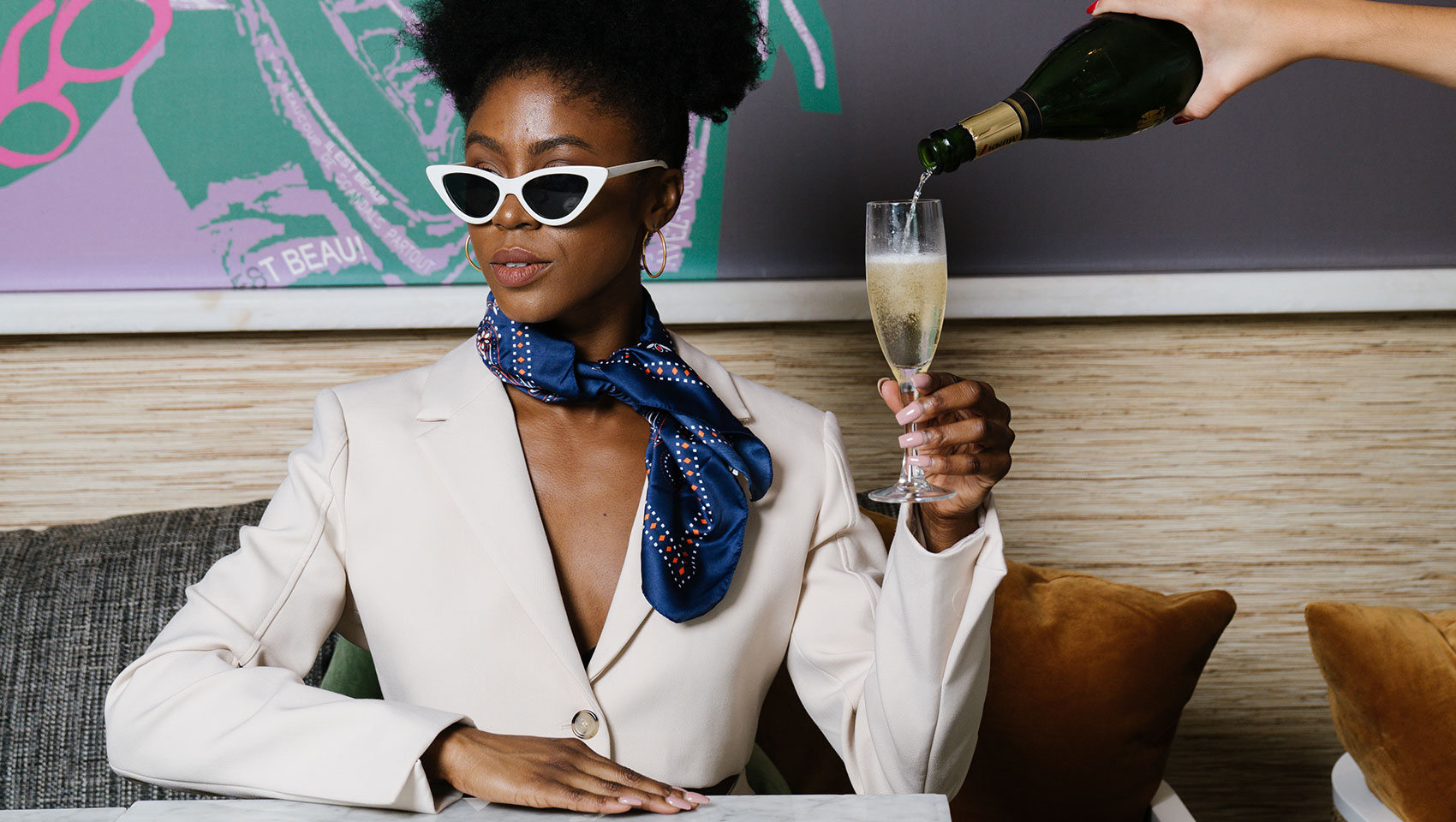 woman sitting at table holding a champagne glass as a disembodied hand fills it with champagne